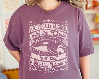 May include: A purple t-shirt with a graphic design featuring a cartoon orca and the text "Politically Aligned with the Orcas who keep sinking Yachts".