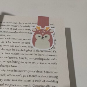 May include: A pink and white deer bookmark with a brown body, white antlers, and a white face. The deer has closed eyes and a small, pink nose.