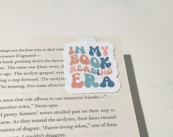 Op de afbeelding: Een vierkante magnetische boekenlegger met de tekst IN MY BOOK READING ERA in een golvend, retro-geïnspireerd lettertype. De letters wisselen af tussen tinten oudroze, lichtblauw en donkerblauw op een effen witte achtergrond.