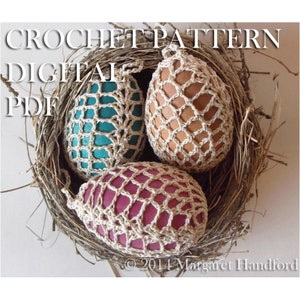 Könnte beinhalten: Drei gehäkelte Eierhüllen in einem Vogelnest. Die Hüllen sind aus beigefarbenem Garn gefertigt und haben ein netzartiges Muster. Die Eier sind türkis, braun und rosa.