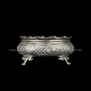 Puede incluir: Un cuenco plateado y ornamentado con un borde festoneado y patrones decorativos. El cuenco descansa sobre tres patas ornamentadas. Las palabras "PEACE" y "JEWELRY IMP" son visibles en los lados del cuenco.