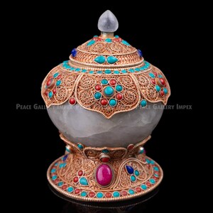 May include: An ornate, handcrafted vessel with a light gray body and a decorative lid. The vessel is adorned with intricate gold filigree, turquoise, and red stone accents. A large pink stone is set on the base. The vessel has a clear, pointed finial.