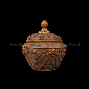 Può includere: Ciotola dorata ornata con coperchio, motivi intricati e abbellimenti. La ciotola presenta pietre rosse e turchesi e perline decorative pendenti. Il coperchio è sormontato da un elemento decorativo. Su sfondo nero.