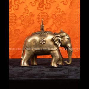 May include: A brass elephant figurine with intricate details, including a decorative saddle and a small figure on its back. The elephant is standing on four legs, with its trunk curled. The background features an orange patterned fabric.