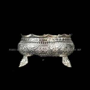 Puede incluir: Un cuenco plateado y ornamentado, con un borde festoneado y tres pies decorativos. El cuenco presenta patrones y diseños en relieve detallados alrededor de su cuerpo, sobre un fondo oscuro.