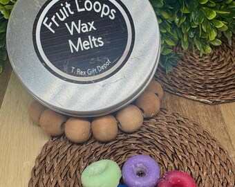 May include: A silver tin with a black label reading "Fruit Loops Wax Melts" sits atop a woven coaster. Below, six colorful, donut-shaped wax melts in green, blue, purple, red, and yellow are arranged on the coaster. The background features greenery.