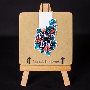 May include: A brown magnetic bookmark with a floral design and the text "Rejoice in the Lord". The bookmark is on a small wooden easel.