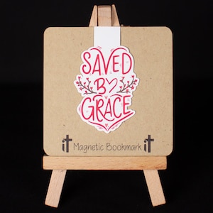 May include: A white magnetic bookmark with the text "SAVED BY GRACE" in red lettering with a heart in the middle of the "BY". The bookmark is on a small wooden easel with the text "it Magnetic Bookmark it" printed on the bottom.