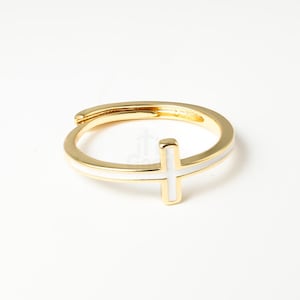 May include: Gold-tone adjustable ring featuring a white enamel cross design. The ring has a polished finish and a simple, elegant style. The cross is centered on the band, creating a minimalist aesthetic.
