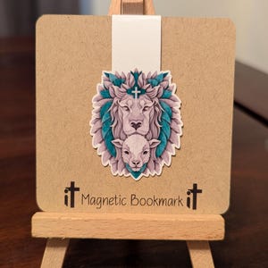 May include: A magnetic bookmark with a white lion with a cross on its head and a white lamb in front of it. The lion and lamb are surrounded by teal leaves. The bookmark is on a brown background with the text "Magnetic Bookmark" in black.