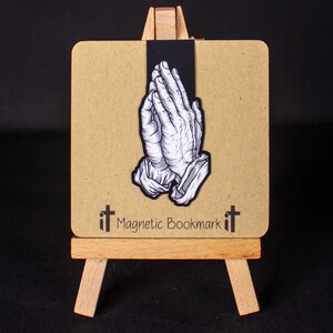 May include: A black and white magnetic bookmark with a design of two hands clasped in prayer. The bookmark is on a small wooden easel and has the text "it Magnetic Bookmark it" printed on it.