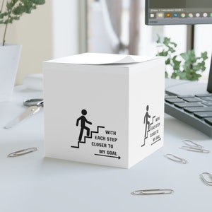 May include: White cube-shaped notepad with a black stick figure walking up stairs. The text on the notepad reads "With each step closer to my goal".