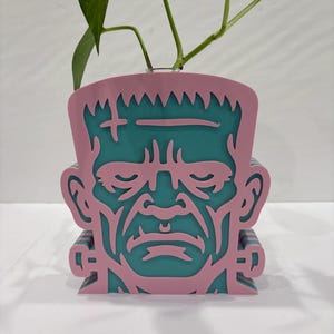 May include: A pink and teal Frankenstein-themed planter. The planter features a detailed cutout design of Frankenstein's face, with a cross and stitches on the forehead. The planter holds a green plant.