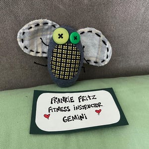 May include: A handmade fabric fly with button eyes and stitched wings. The fly has a patterned body and is paired with a card that reads "FRANKIE FRITZ FITNESS INSTRUCTOR GEMINI".