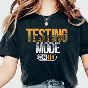 May include: Black t-shirt with the text "TESTING MODE ON" in yellow and white. The shirt is worn with blue jeans and white sneakers. The shirt's design features a switch graphic.
