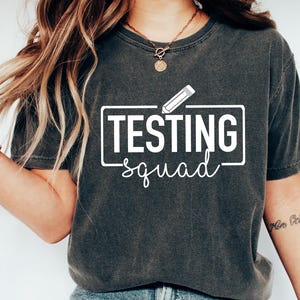 May include: Dark gray t-shirt with the text "TESTING squad" in a white rectangular box with a pencil graphic. The shirt is paired with blue jeans and white sneakers. The overall style is casual and comfortable.