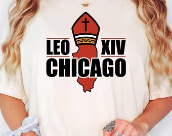 Könnte beinhalten: Cremefarbenes T-Shirt mit einem grafischen Design. Das Design enthält den Text "LEO XIV CHICAGO" in Schwarz, mit einer roten und schwarzen Illustration einer Bischofsmütze und des Bundesstaates Illinois. Das Shirt ist ein lässiger Stil.