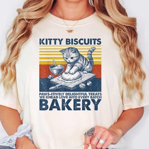 May include: Cream-colored t-shirt featuring a graphic of a cat kneading dough, with text that reads "Kitty Biscuits" and "Bakery." The design includes a retro color scheme of orange, yellow, and blue.