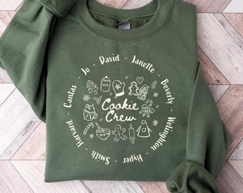 May include: An olive green sweatshirt with the words "Cookie Crew" in a circular design. The design includes illustrations of baking tools and ingredients, with names around the circle. The sweatshirt is made of a soft material.