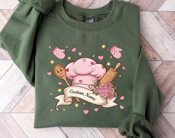 Op de afbeelding: Groen sweatshirt met een chef-thema ontwerp. Het ontwerp bevat een roze koksmuts, deegroller, houten lepel en roze harten. De tekst "Custom Name" staat op een banner. Twee roze ovenwanten en sterren zijn ook afgebeeld.