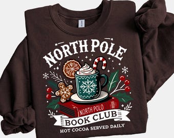 May include: A brown sweatshirt with a graphic that reads "North Pole Book Club" with a hot cocoa mug, gingerbread man, and candy cane design. The sweatshirt is a crew neck style.