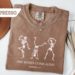 May include: Espresso-colored t-shirt with a white skeleton graphic. The shirt features the text "DRY BONES COME ALIVE" and "Ezekiel 37". A silver necklace with a pendant is draped on the shirt.