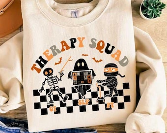 May include: A cream-colored sweatshirt with the text "Therapy Squad" in orange and black letters. The text is surrounded by a black and white checkered pattern. There are three Halloween-themed characters in the center of the design: a skeleton, a ghost, and a mummy.