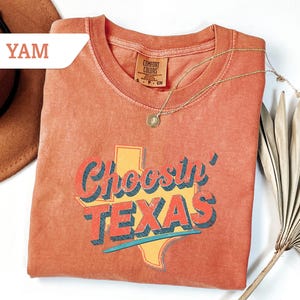 May include: A coral-colored t-shirt with the phrase "Choosin' TEXAS" in a retro font, featuring a yellow outline of the state. A gold necklace with a pendant rests on the shirt. The shirt is folded, and a brown hat is partially visible.