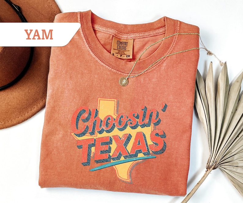 May include: A coral-colored t-shirt with the phrase "Choosin' TEXAS" in a retro font, featuring a yellow outline of the state. A gold necklace with a pendant rests on the shirt. The shirt is folded, and a brown hat is partially visible.