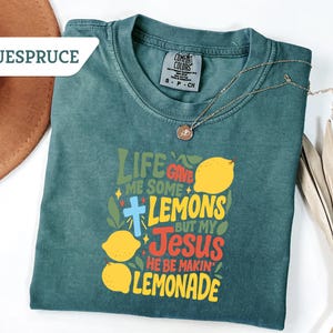 May include: A teal Comfort Colors t-shirt with the text "Life Gave Me Some Lemons But My Jesus He Be Makin' Lemonade" in yellow, red, and green. A gold necklace with a pendant is draped across the shirt.