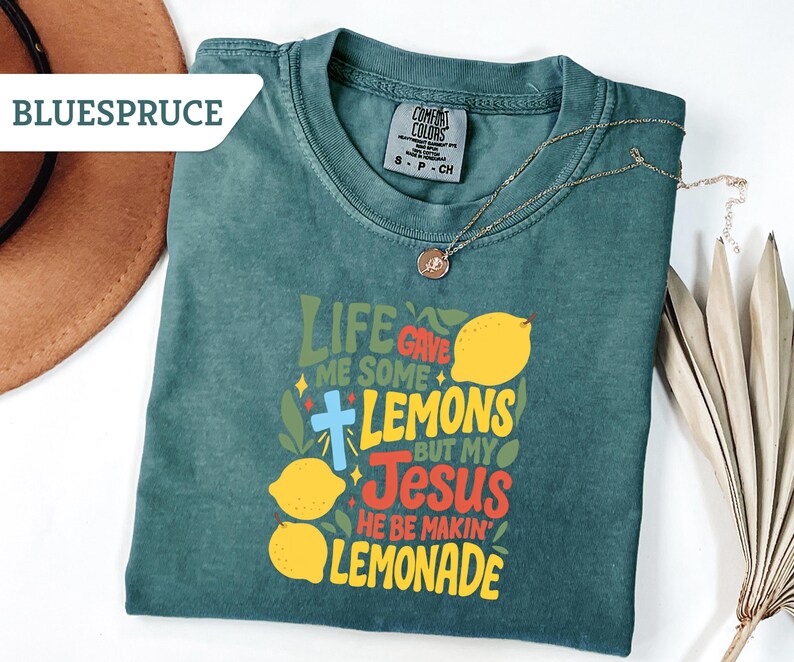 May include: A teal Comfort Colors t-shirt with the text "Life Gave Me Some Lemons But My Jesus He Be Makin' Lemonade" in yellow, red, and green. A gold necklace with a pendant is draped across the shirt.