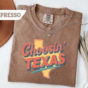 May include: Espresso-colored t-shirt with the text "Choosin' TEXAS" in red, orange, and blue, over a yellow Texas outline. A silver necklace with a pendant is draped on the shirt. A brown hat is visible in the upper left corner.