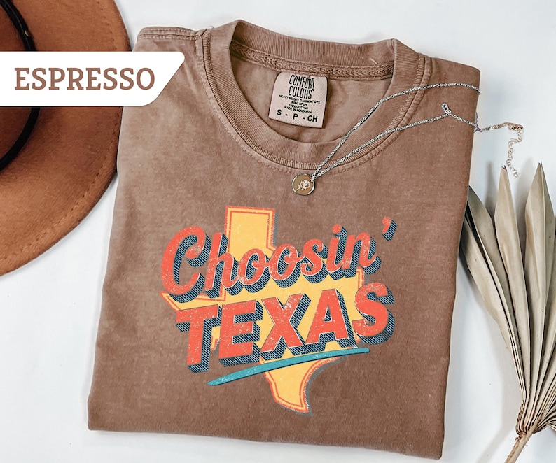 May include: Espresso-colored t-shirt with the text "Choosin' TEXAS" in red, orange, and blue, over a yellow Texas outline. A silver necklace with a pendant is draped on the shirt. A brown hat is visible in the upper left corner.