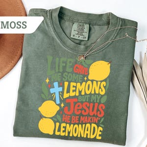 May include: Moss green t-shirt with the text "Life Gave Me Some Lemons But My Jesus He Be Makin' Lemonade" in colorful lettering, featuring lemon illustrations and a cross. A gold necklace and a brown hat are also visible.