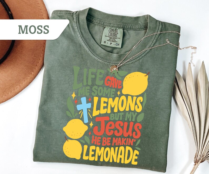 May include: Moss green t-shirt with the text "Life Gave Me Some Lemons But My Jesus He Be Makin' Lemonade" in colorful lettering, featuring lemon illustrations and a cross. A gold necklace and a brown hat are also visible.