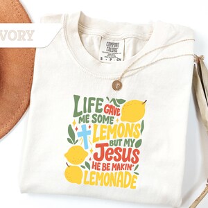 May include: Ivory-colored t-shirt with the text "LIFE GAVE ME SOME LEMONS BUT MY JESUS HE BE MAKIN' LEMONADE" in green, yellow, and red. The design includes lemons and a cross. A gold necklace is draped across the shirt.