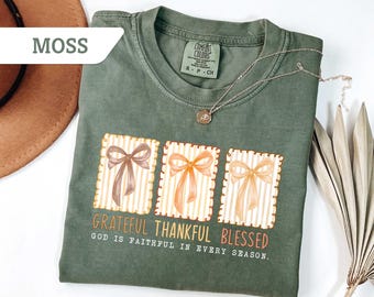 May include: Moss green Comfort Colors t-shirt with a graphic of three gift boxes with bows. The text "Grateful Thankful Blessed" and "God is faithful in every season" are printed below the boxes. A silver necklace with a pendant is draped on the shirt.
