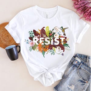 May include: White t-shirt with a floral design and the word "RESIST" printed in black. The design features a hummingbird, leaves, and flowers in shades of red, orange, yellow, and green.