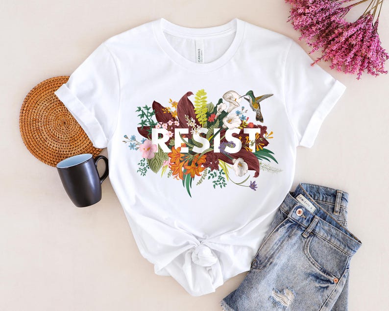 May include: White t-shirt with a floral design and the word "RESIST" printed in black. The design features a hummingbird, leaves, and flowers in shades of red, orange, yellow, and green.