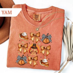 May include: A coral-colored sweatshirt featuring a Thanksgiving-themed graphic. The design includes images of pumpkins with checkered bows, pie slices, and a turkey wearing a pilgrim hat. The word "YAM" is in a white banner in the upper left corner.