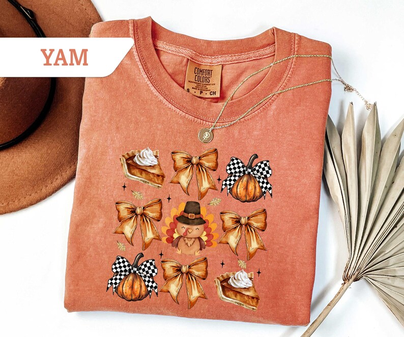 May include: A coral-colored sweatshirt featuring a Thanksgiving-themed graphic. The design includes images of pumpkins with checkered bows, pie slices, and a turkey wearing a pilgrim hat. The word "YAM" is in a white banner in the upper left corner.