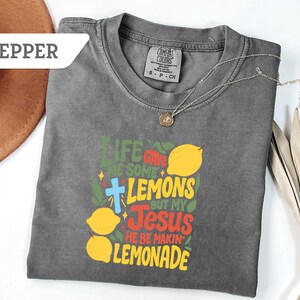 May include: A gray Comfort Colors t-shirt with the text "Life Gave Me Some Lemons But My Jesus He Be Makin' Lemonade" in green, yellow, red, and blue. The shirt features lemon graphics and a cross. A silver necklace with a pendant is on the shirt.