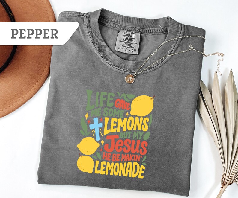 May include: A gray Comfort Colors t-shirt with the text "Life Gave Me Some Lemons But My Jesus He Be Makin' Lemonade" in green, yellow, red, and blue. The shirt features lemon graphics and a cross. A silver necklace with a pendant is on the shirt.