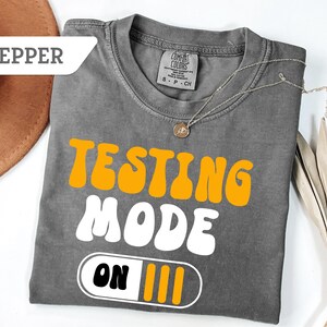 May include: A gray t-shirt with the words "TESTING MODE ON" in orange and white lettering. The shirt has a crew neck and short sleeves. A gold necklace with a pendant is draped on the shirt.