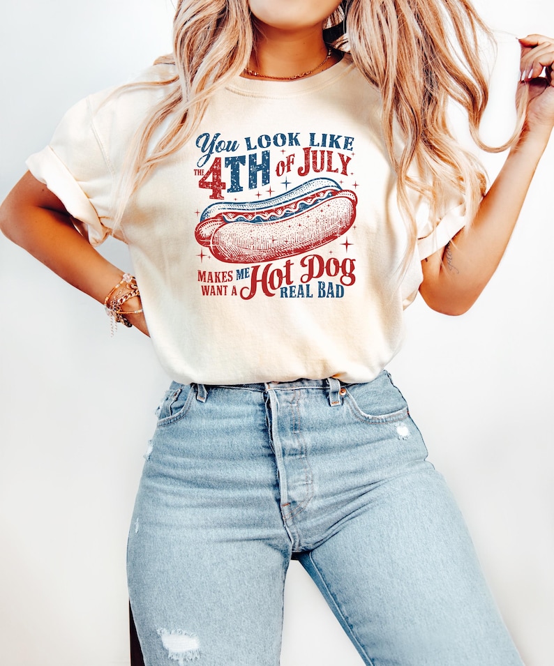 Op de afbeelding: Cr&egrave;mekleurig T-shirt met een afbeelding van een hotdog en de tekst "You look like the 4th of July makes me want a hot dog real bad." Het ontwerp bevat rood, wit en blauw.