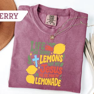 May include: Berry-colored t-shirt with the text "Life gave me some lemons but my Jesus he be makin' lemonade" in green, yellow, and red. Features a cross and lemon graphics. A gold necklace is draped across the shirt.