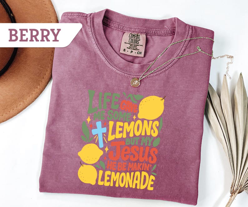 May include: Berry-colored t-shirt with the text "Life gave me some lemons but my Jesus he be makin' lemonade" in green, yellow, and red. Features a cross and lemon graphics. A gold necklace is draped across the shirt.