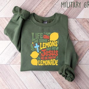 May include: Military green sweatshirt with the text "Life gave me some lemons but my Jesus he be makin' lemonade" in yellow, red, and green. The design includes lemons and a cross. The sweatshirt is laid flat on a light wood grain surface.