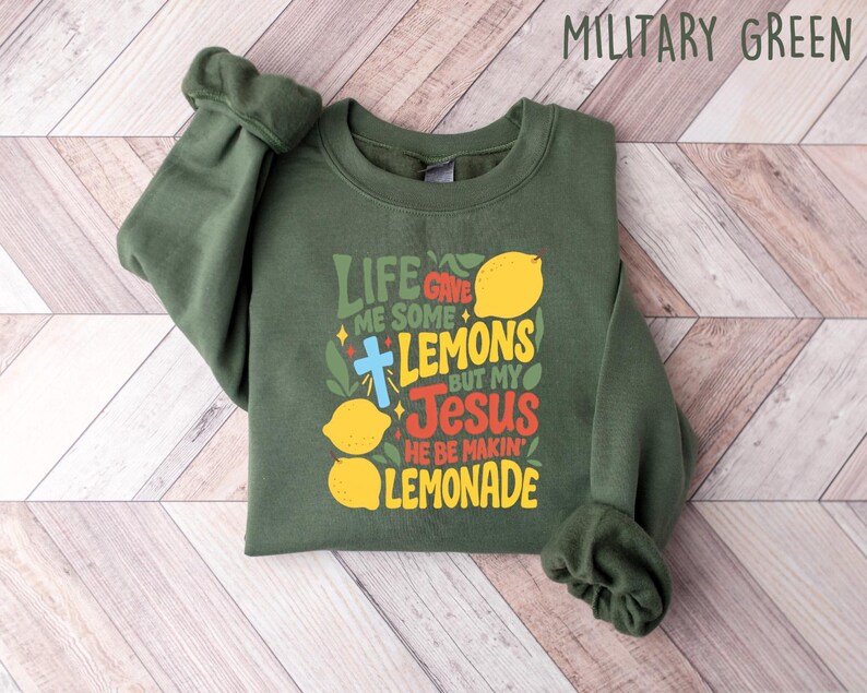 May include: Military green sweatshirt with the text "Life gave me some lemons but my Jesus he be makin' lemonade" in yellow, red, and green. The design includes lemons and a cross. The sweatshirt is laid flat on a light wood grain surface.