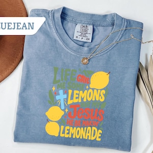 May include: Bluejean colored t-shirt with the text "Life Gave Me Some Lemons, So I Made Jesus Lemonade" in green, yellow, and red. The shirt features a cross and lemon graphics. A gold necklace and a brown hat are also visible.
